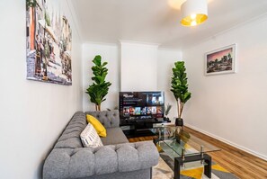 Ein modernes Wohnzimmer mit grauem Sofa, einem Glastisch, einem Flachbildfernseher und zwei Topfpflanzen.