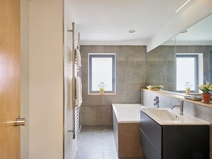 A modern bathroom with a bathtub, a sink, and a large mirror. There are two windows providing natural light. - The Chatterbox at Waterloo Farm (Halesworth)