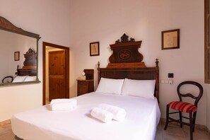 A neatly made bed with white linens and two rolled towels, a wooden nightstand with a lamp, a wooden chair with a red cushion, and a wooden headboard with a decorative crest. - Son Idò by Rentallorca (Muro)
