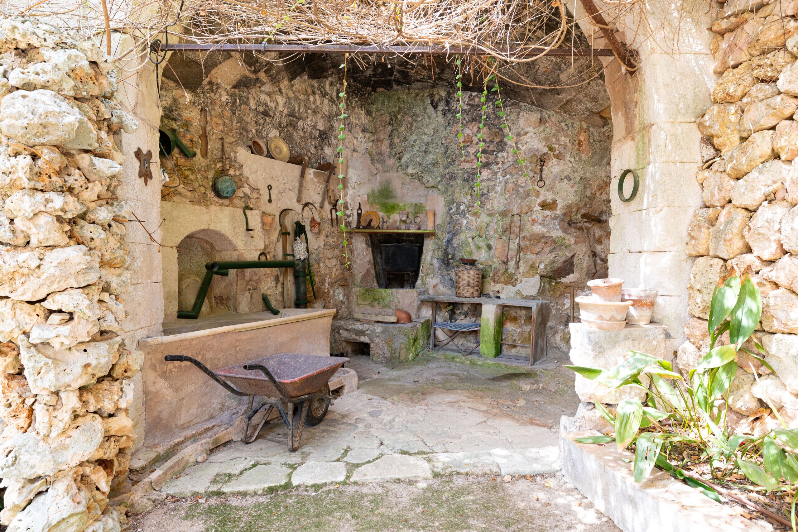 Une cour rustique aux murs de pierre, agrémentée d’une brouette, d’une plante suspendue, ainsi que de divers outils et poteries en pierre.