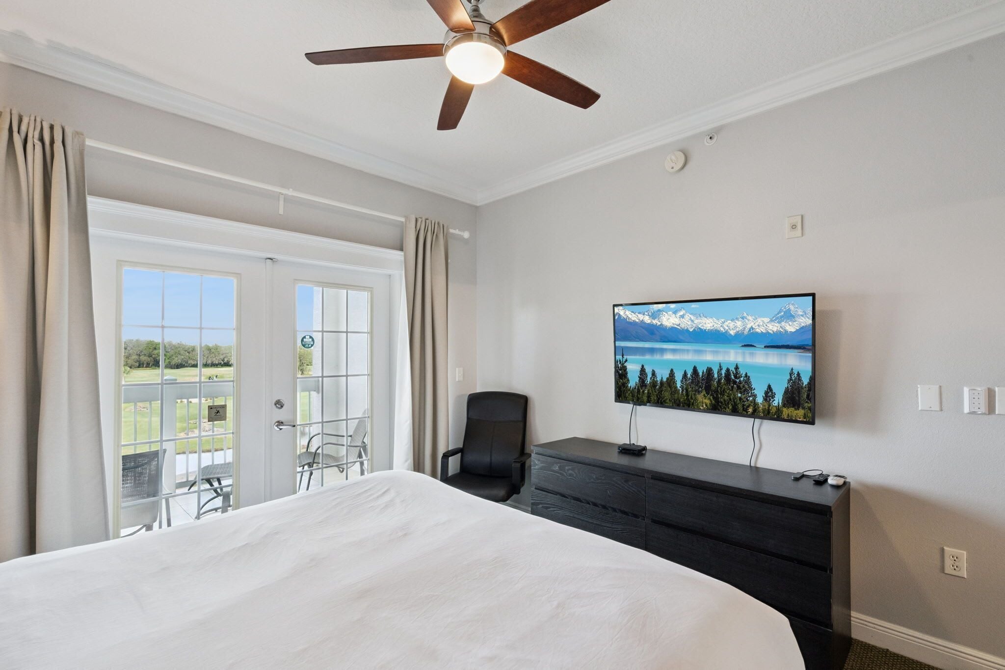 A hotel room with a large bed, a ceiling fan, a television on the wall, and a view of a mountain landscape through the window.