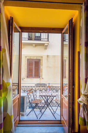 Ein offener Balkon mit Tisch und Stühlen, ein Fenster mit Rollläden und Blick auf ein Gebäude gegenüber.