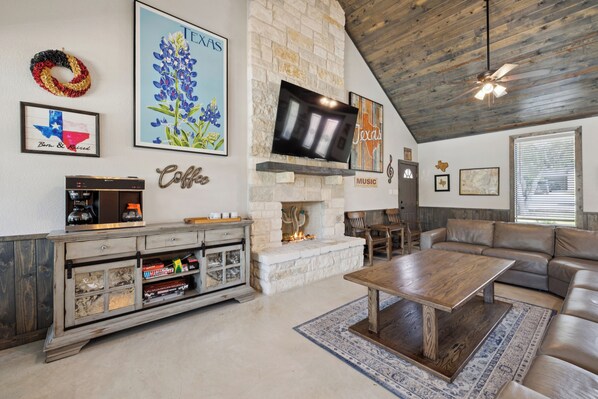 A spacious living room with a stone fireplace, a flat-screen TV mounted on the wall, a coffee bar, and a leather sofa.