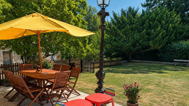 Una veranda esterna con un tavolo e sedie in legno, un ombrello giallo e un lampione.