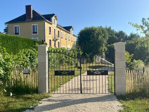 Una entrada tancada amb reixa a un edifici groc amb un camí de grava, tot envoltat de vegetació.