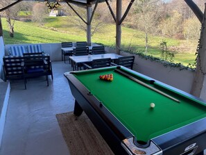 Une table de billard avec des billes, une queue de billard et un tapis vert. Des chaises et des tables sont disposées autour de la table de billard, et on aperçoit un espace verdoyant avec des arbres en arrière-plan.