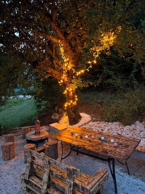 An outdoor seating area with a wooden table and benches, surrounded by a tree with string lights.
