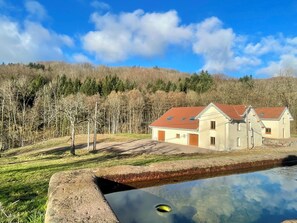 Un edifici de dues plantes amb teulada vermella, piscina i un turó boscós al fons.
