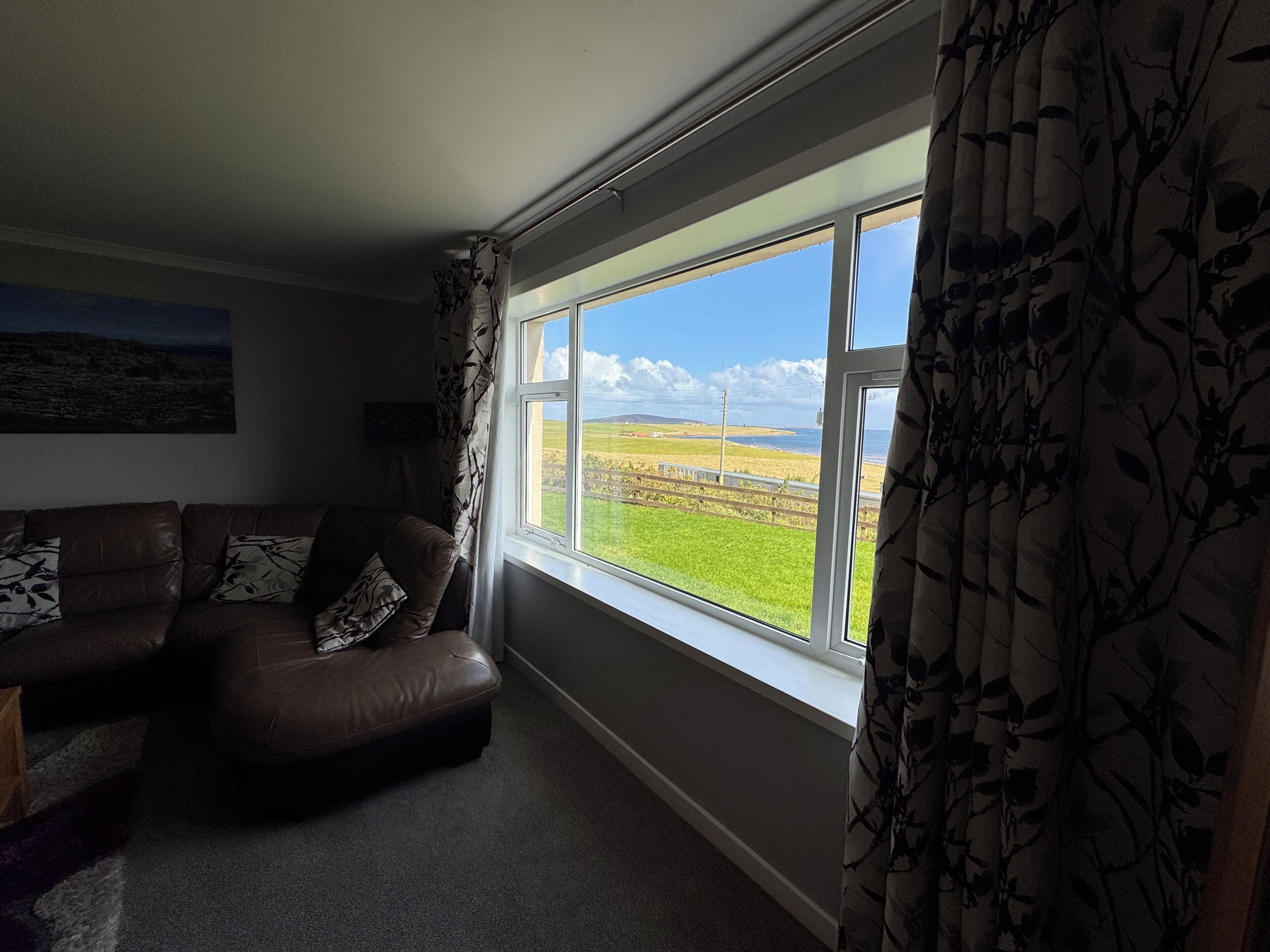 A living room with a brown leather sofa, a window with a view of a green field and a clear sky, and a patterned curtain.