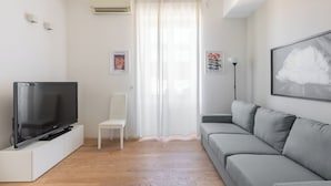 A living room with a flat-screen TV on a white stand, a grey sofa, a white chair, and a large window with sheer curtains. - San Gervasio Apartment by Wonderful Italy (Bologna)