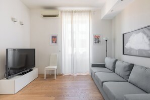 Ein Wohnzimmer mit einem Flachbildfernseher auf einem weißen Schrank, einem grauen Sofa, einem weißen Sessel und einem großen Fenster mit leichtem Vorhang.