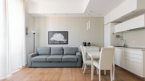 A modern living space with a grey sofa, white dining table, and chairs, a kitchen area with white cabinets, and a framed picture on the wall. - San Gervasio Apartment by Wonderful Italy (Bologna)