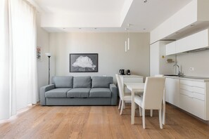 Ein moderner Wohnraum mit grauem Sofa, weißem Esstisch und Stühlen, einer Küchenzeile mit weißen Schränken sowie einem gerahmten Bild an der Wand.