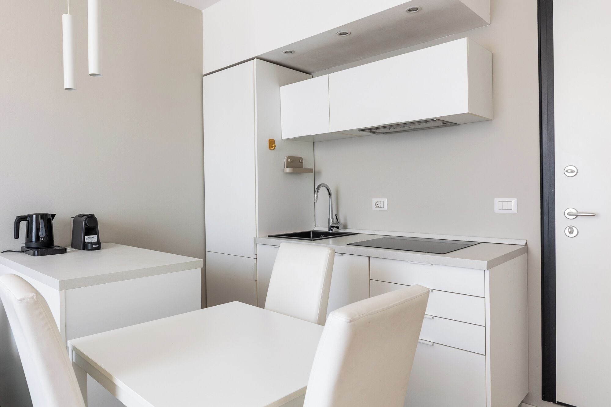 A modern kitchenette with a sink, stove, and white cabinets. There is a table with two chairs and a coffee maker on the counter.