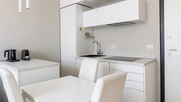 A modern kitchenette with a sink, stove, and white cabinets. There is a table with two chairs and a coffee maker on the counter. - San Gervasio Apartment by Wonderful Italy (Bologna)