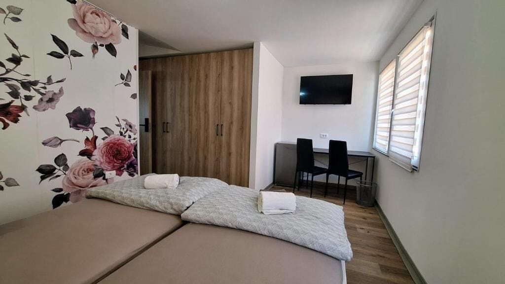 Ein Zimmer mit einem Bett, einem Fernseher, einem Fenster mit Jalousien und einer Tür mit floralem Wandpapier.