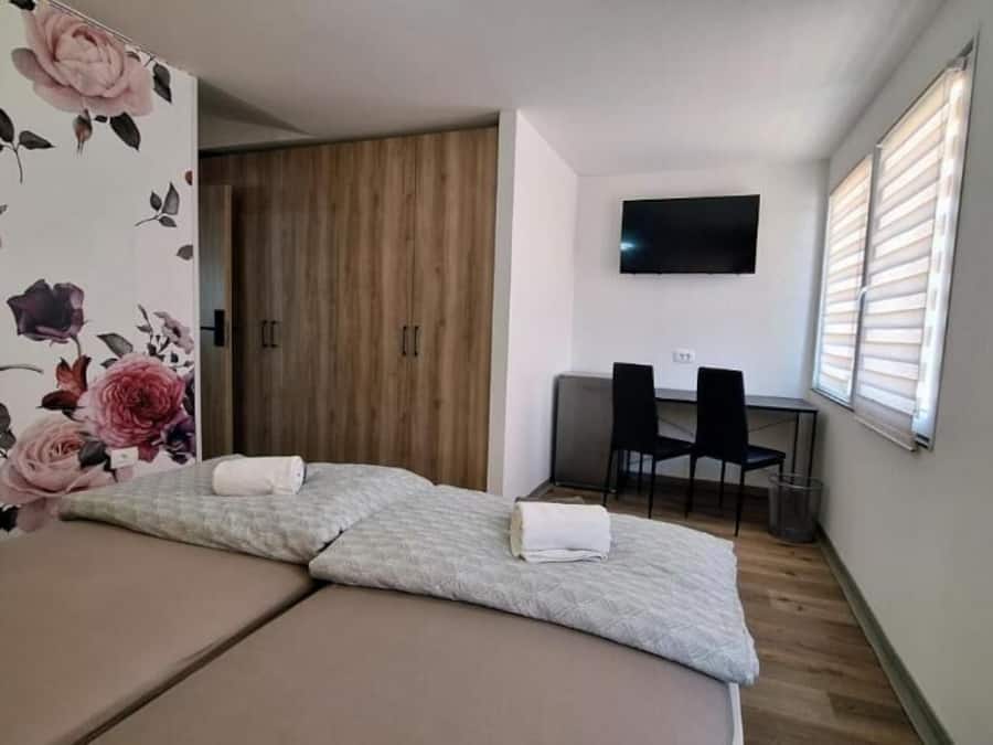 Una habitación con cama, televisor, ventana con persianas y puerta con papel tapiz floral.