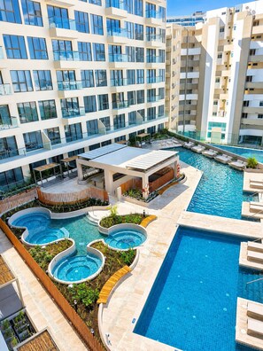 Un hotel de varios pisos con un área de piscina en la azotea, que incluye una piscina central grande y piscinas circulares más pequeñas.