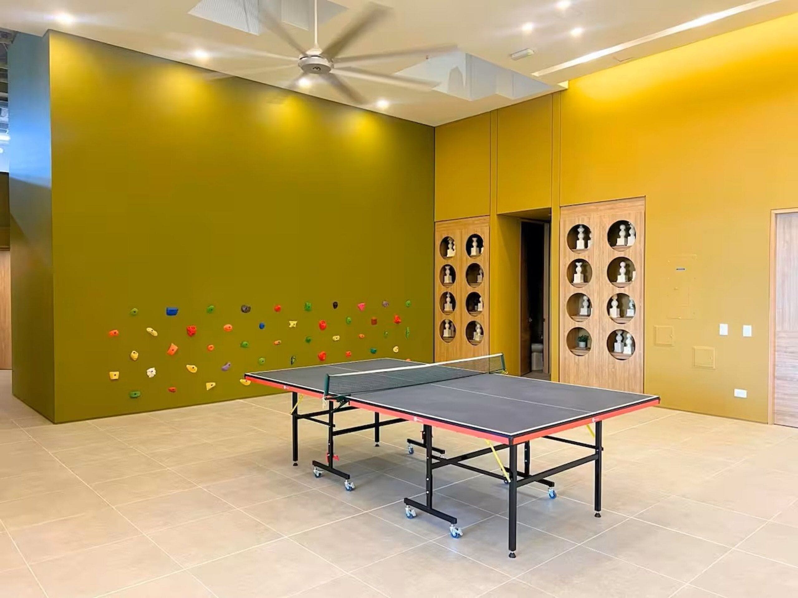 Una mesa de ping pong en una habitación con una pared de escalada y un ventilador de techo.