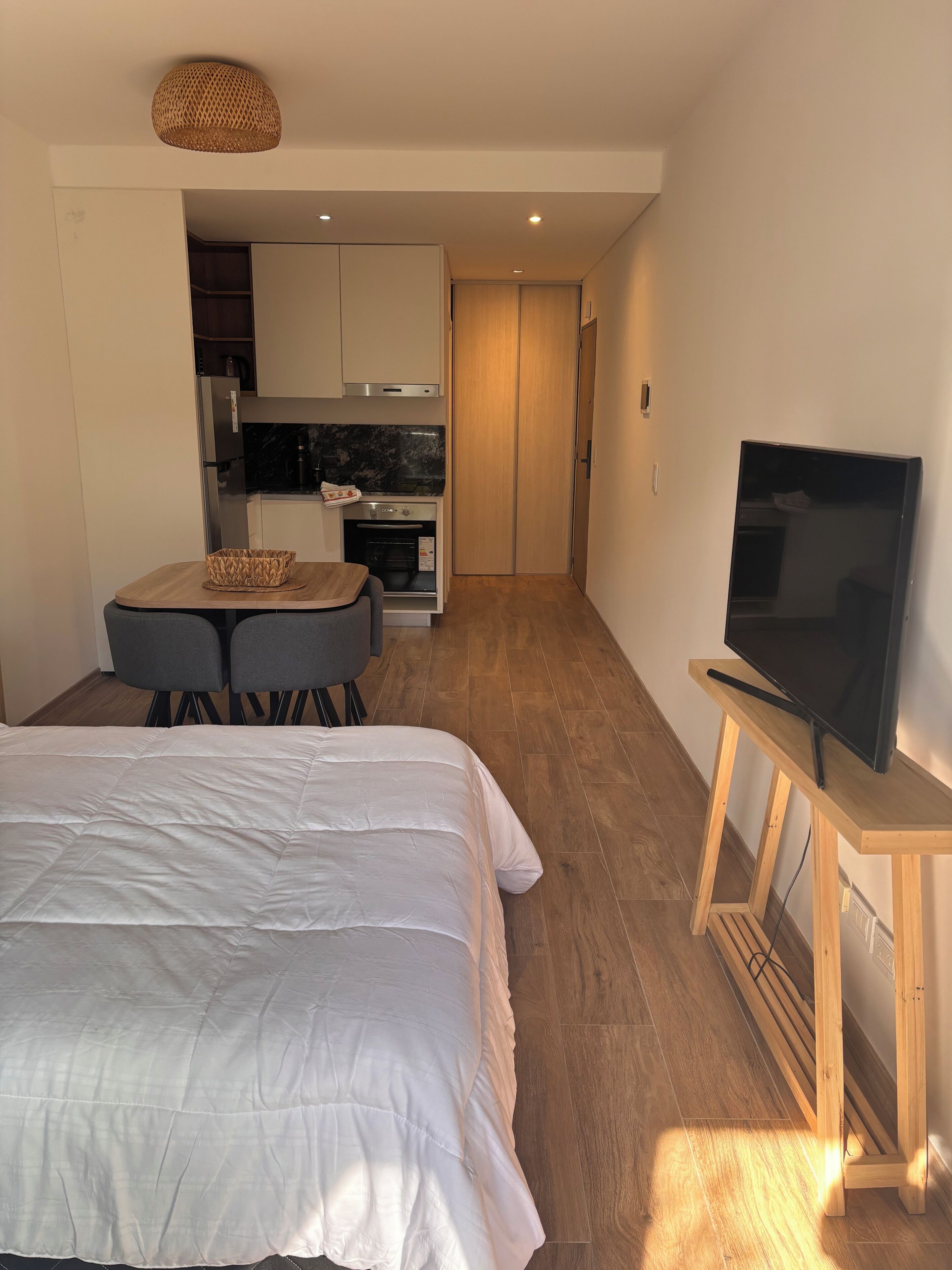 Un dormitorio con cama, televisor sobre un mueble, kitchenette con estufa y horno, y zona de comedor con mesa y sillas.