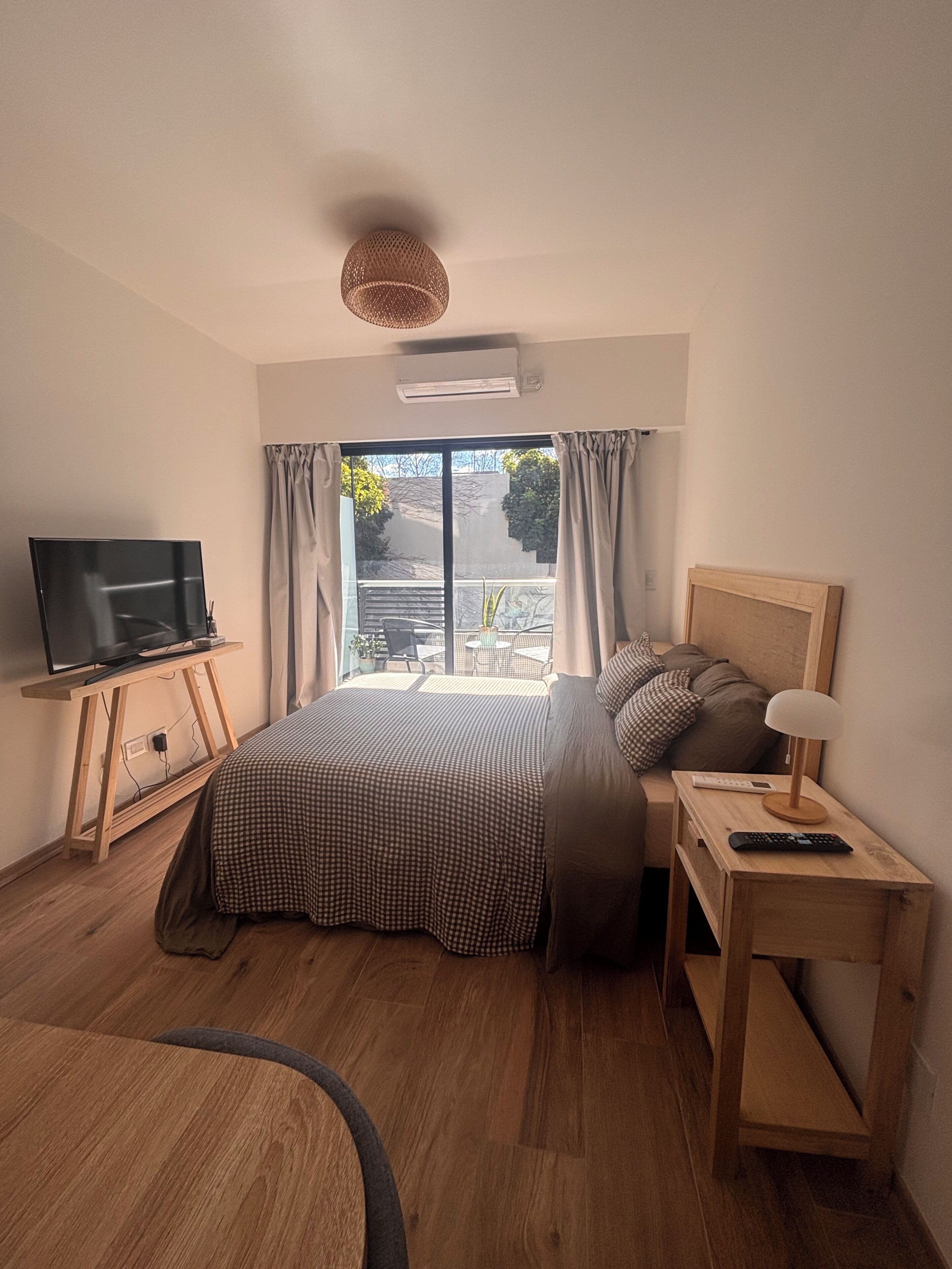 Une chambre à coucher avec un lit, un téléviseur posé sur un meuble en bois, une table de chevet avec une lampe et une fenêtre avec des rideaux.
