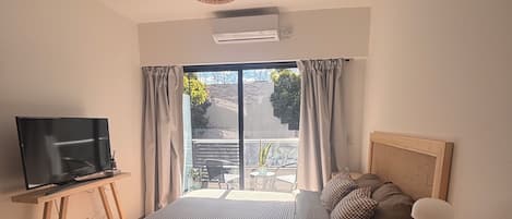A bedroom with a bed, a TV on a wooden stand, a bedside table with a lamp, and a window with curtains.