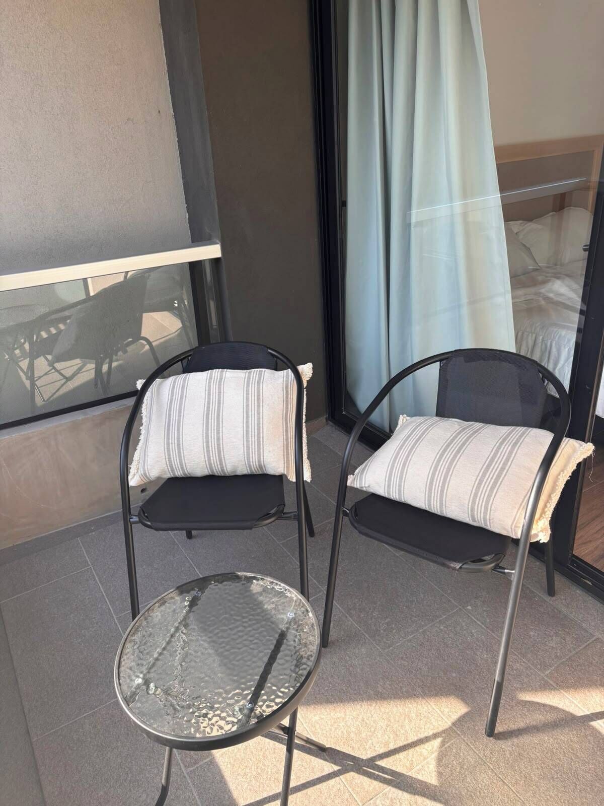 Un balcón con dos sillas, una mesita pequeña, una puerta de cristal y vista a una cama a través de las cortinas.