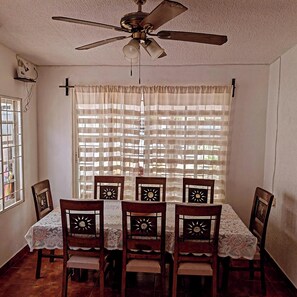 A dining area with a table set for four, wooden chairs with intricate back designs, a ceiling fan, and a window with a view of buildings. - Casa Sol Bacocho (Puerto Escondido)
