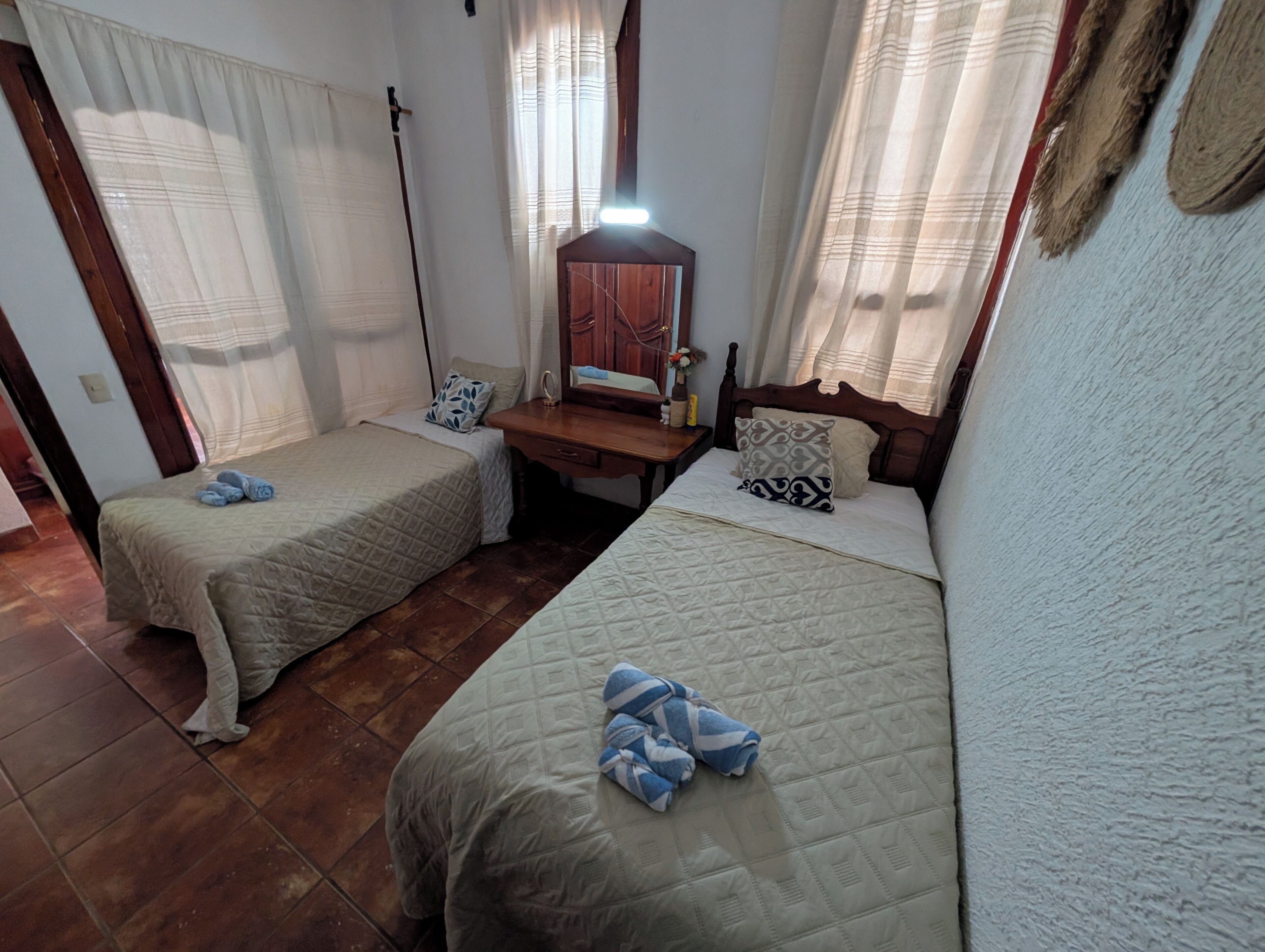 Une chambre avec deux lits, une table de chevet en bois, un miroir et une fenêtre avec des rideaux.