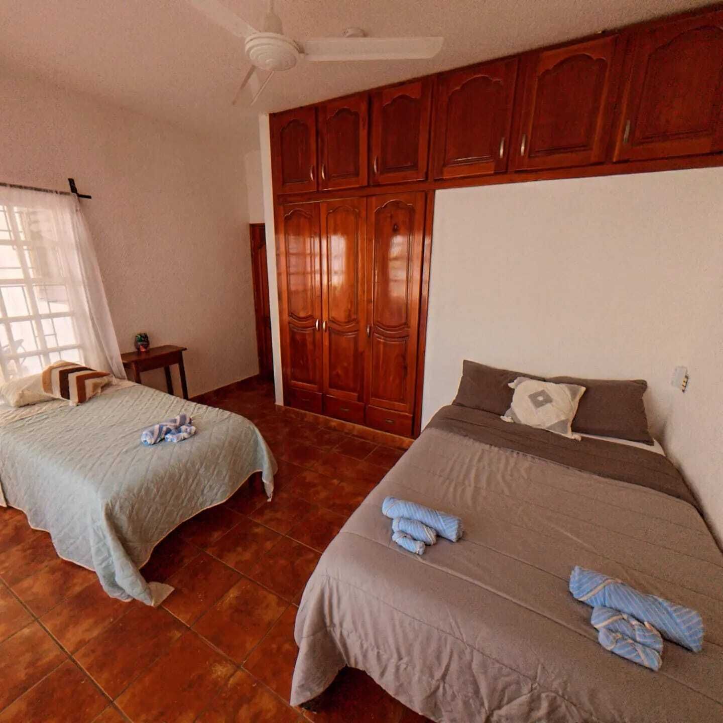 A room with two beds, a ceiling fan, wooden wardrobes, and a tiled floor.