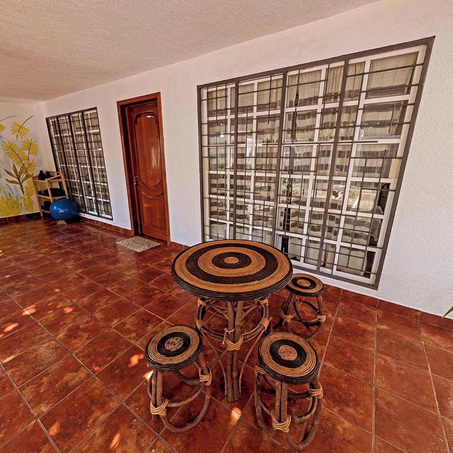 A room with a tiled floor, a wooden door, a large window with a grid pattern, and a round table with stools.