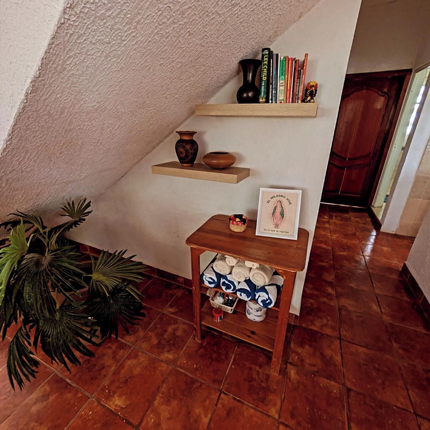 A corner with a shelf holding books and a small vase, a wooden table with towels, and a potted plant.