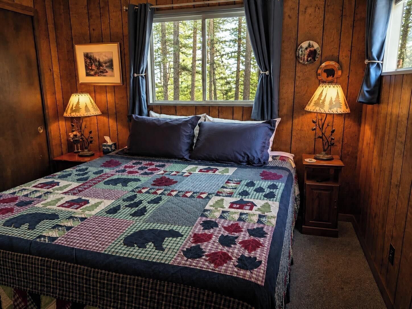 Una habitación de madera con una cama, dos lámparas de noche, un ventanal con cortinas y un cuadro colgado en la pared.