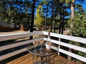 Een glazen tafel met twee wijnglazen en een fles wijn op een houten terras met een witte balustrade, met uitzicht op een bebost gebied.