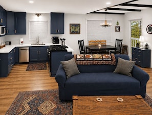 Eine moderne Küche mit dunkelblauen Schränken, ein Essbereich mit Tisch und Stühlen und ein blaues Sofa mit einem Zierkissen.