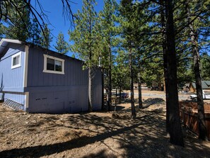 A blue cabin with a small window, surrounded by pine trees and a dirt ground. - Big Bear Escape: Cozy Fireplace! Dine Al Fresco! (Big Bear City)