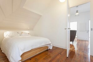 Une chambre avec un plancher en bois, un lit blanc et une porte qui donne sur une autre pièce.