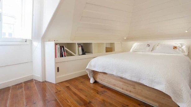 Een slaapkamer met een schuine plafond, een bed met witte beddengoed, houten vloeren en ingebouwde planken met boeken.