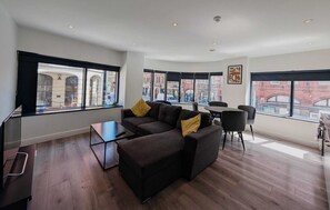 Ein modernes Wohnzimmer mit einer Couch, einem Couchtisch und einem Essbereich. Große Fenster bieten Blick auf die Stadt.