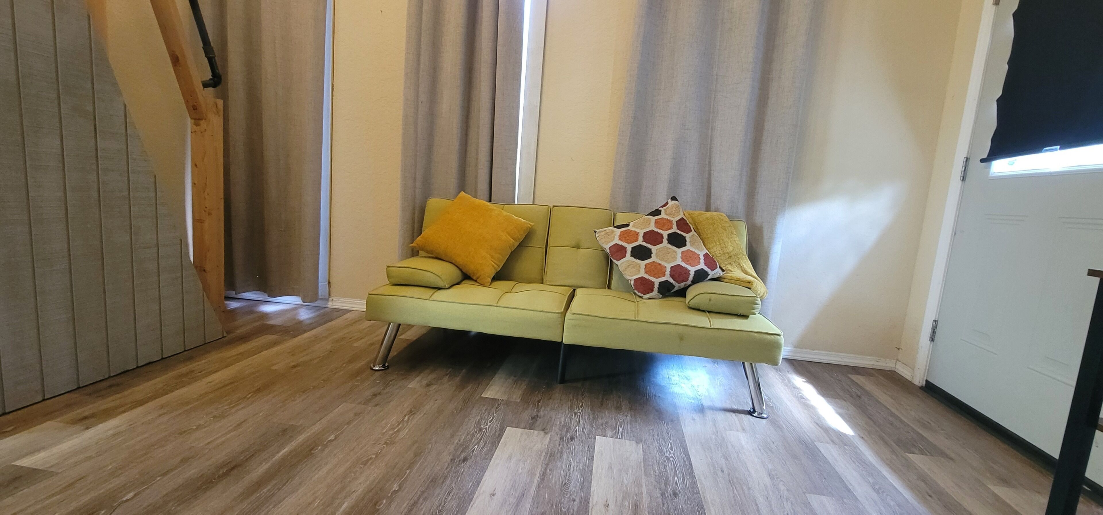 Una habitación con un sofá verde, piso de madera y una ventana con cortinas.