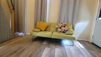 Una habitación con un sofá verde, piso de madera y una ventana con cortinas.
