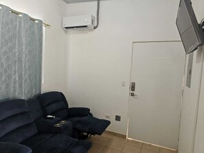 Ein Zimmer mit einem dunkelblauen Sofa, einer Klimaanlage, einem an der Wand befestigten Fernseher und einer Tür.