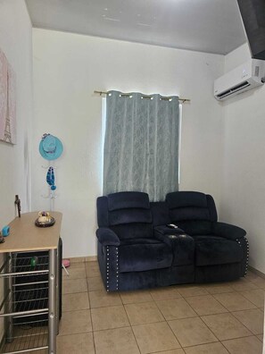 Ein Wohnzimmer mit einem dunkelblauen Sofa, einem kleinen Tisch mit einem dekorativen Gegenstand, einem Fenster mit Vorhängen und einer Klimaanlage an der Wand.