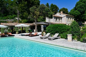A swimming pool with lounge chairs and umbrellas, surrounded by greenery and a building with a tiled roof. - Le Domaine des Oliviers · 6 BR · private pool (Tarascon)