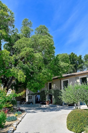Una propietat residencial amb un carrer pavimentat, un edifici de dues plantes amb teulada de rajola, i diversa vegetació com ara arbres i arbustos.