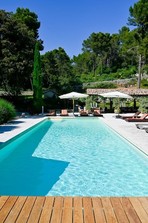 Una piscina amb gandules i ombrel·les, envoltada d'arbres i una paret de pedra.