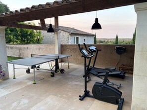 Eine Terrasse im Freien mit einem Tischtennisplatte, Fitnessgeräten und einer Pergola mit hängenden Lichtern.