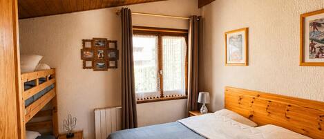 A bedroom with a bunk bed, a single bed, a window with curtains, a bedside table with a lamp, and framed pictures on the wall.
