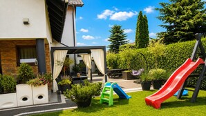 Et beboelsesområde udendørs med terrasse, siddepladser, rutsjebane og pergola.