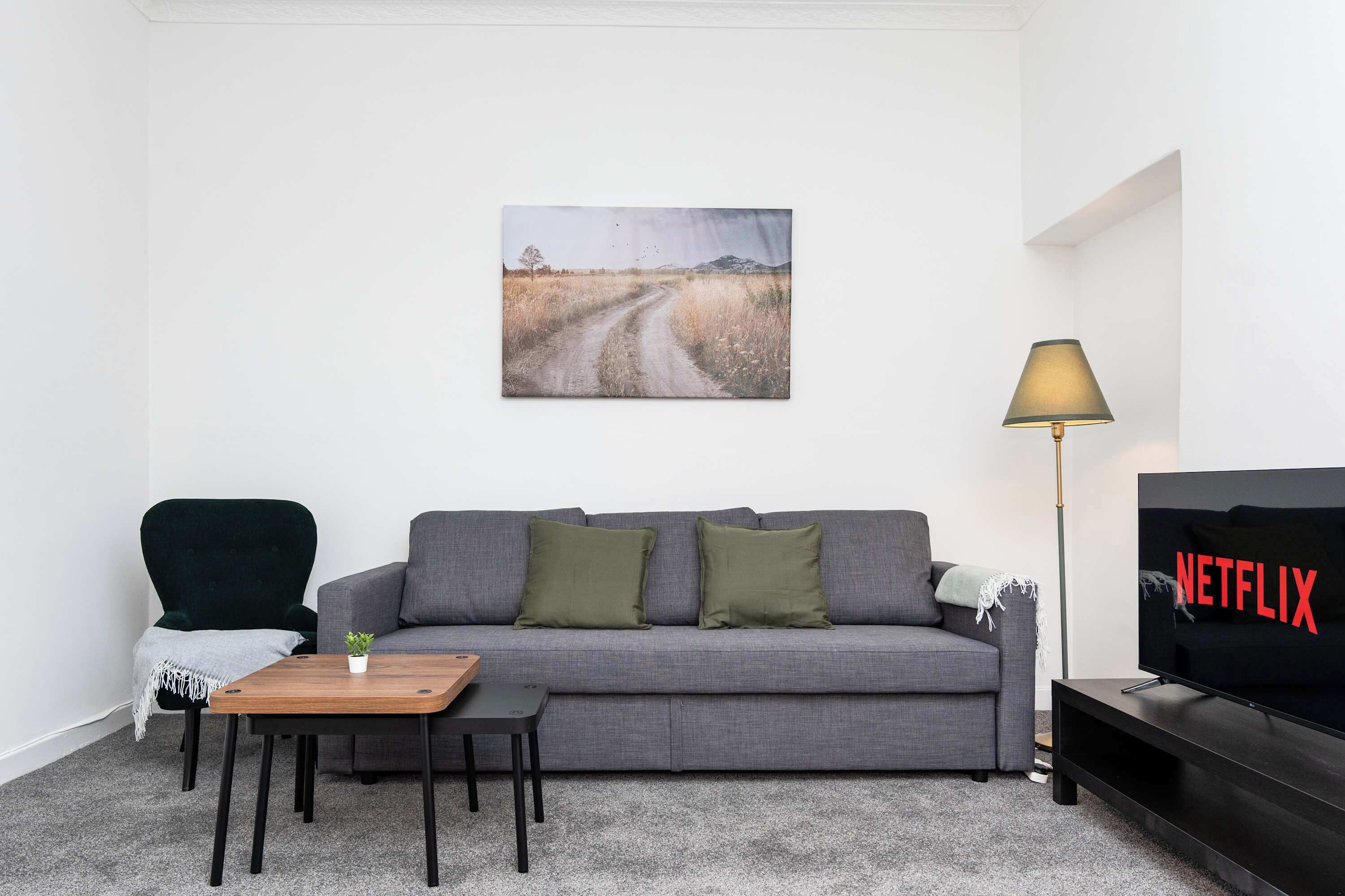 Un salon avec un canapé gris, une petite table en bois, un fauteuil vert, un lampadaire et un téléviseur affichant le logo de Netflix.